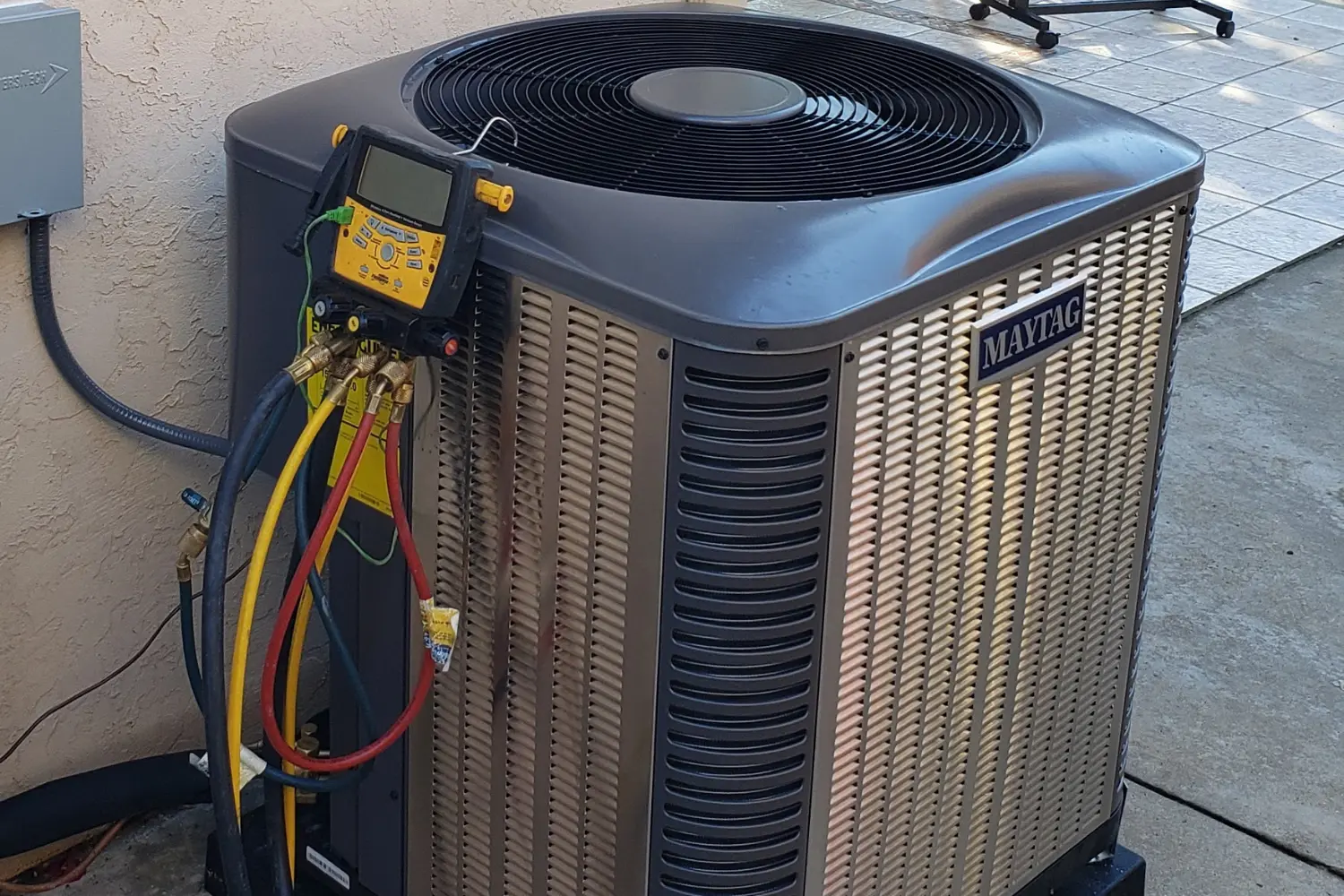 A close up view shows a gray Maytag air conditioning unit with various colored hoses and gauges attached to its side, sitting on a concrete patio next to a stucco wall